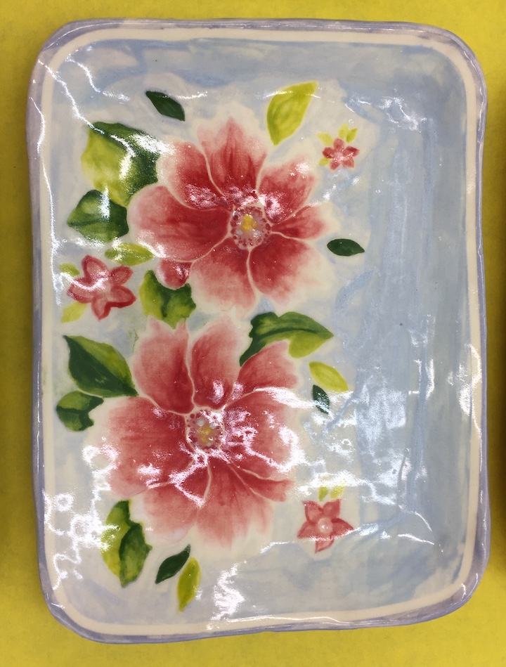 a rectangular ceramic plate in blue, with two overlapping red flowers whose petals gradiates to white at the edges. The flowers have leaves painted around them. The plate is glazed.