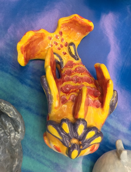 a clay sculpture of a goldfish, with stylized fins and body. The fish is painted yellow and red, with details in purple. The ceramic is glazed.