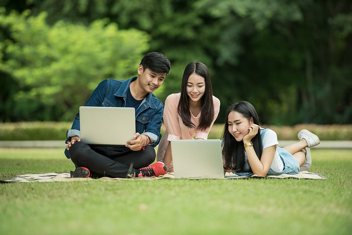 three people with computers and books around them sitting on a grass field